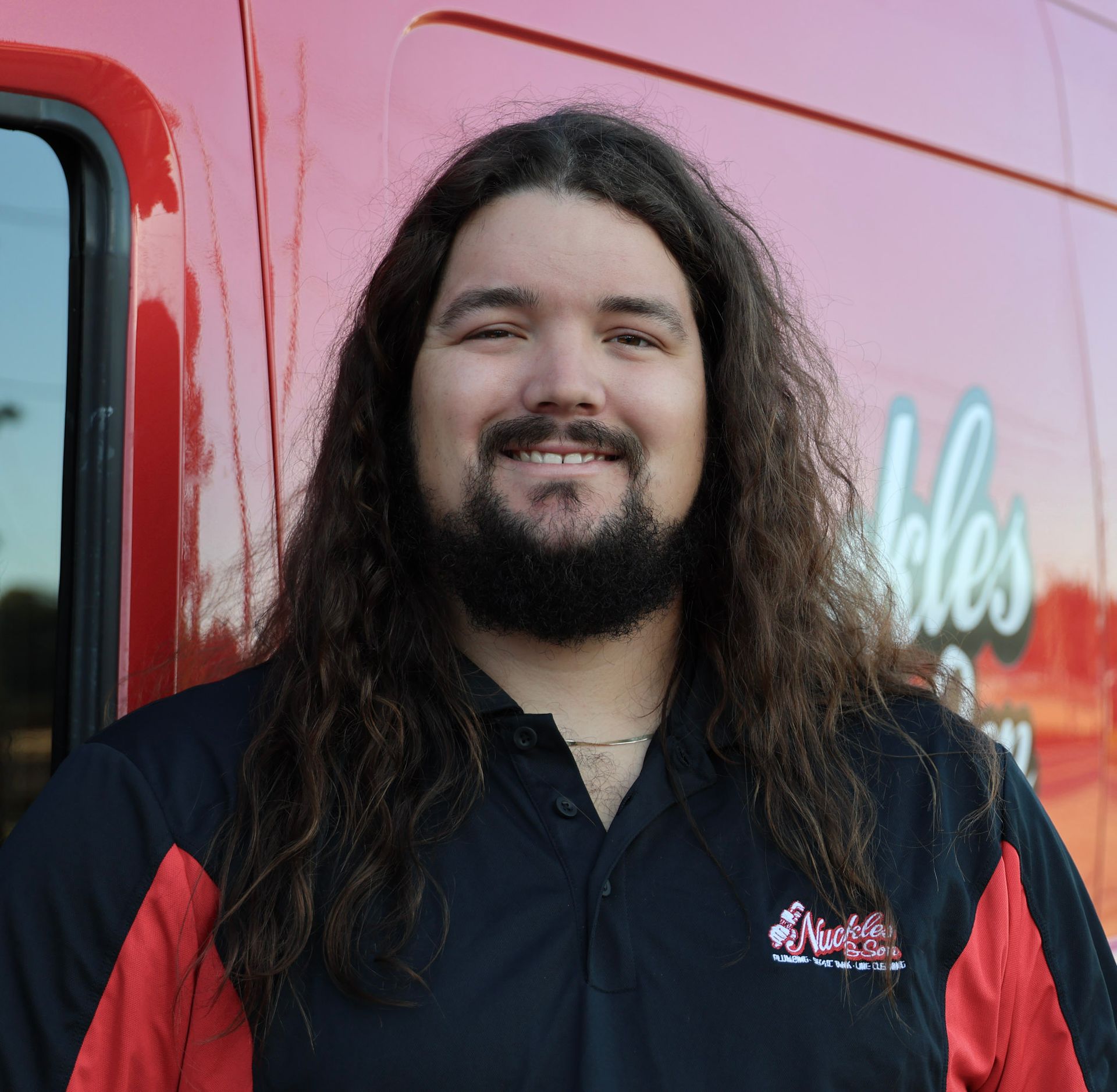 Smiling person with long dark hair in a black-and-red shirt standing beside a red vehicle