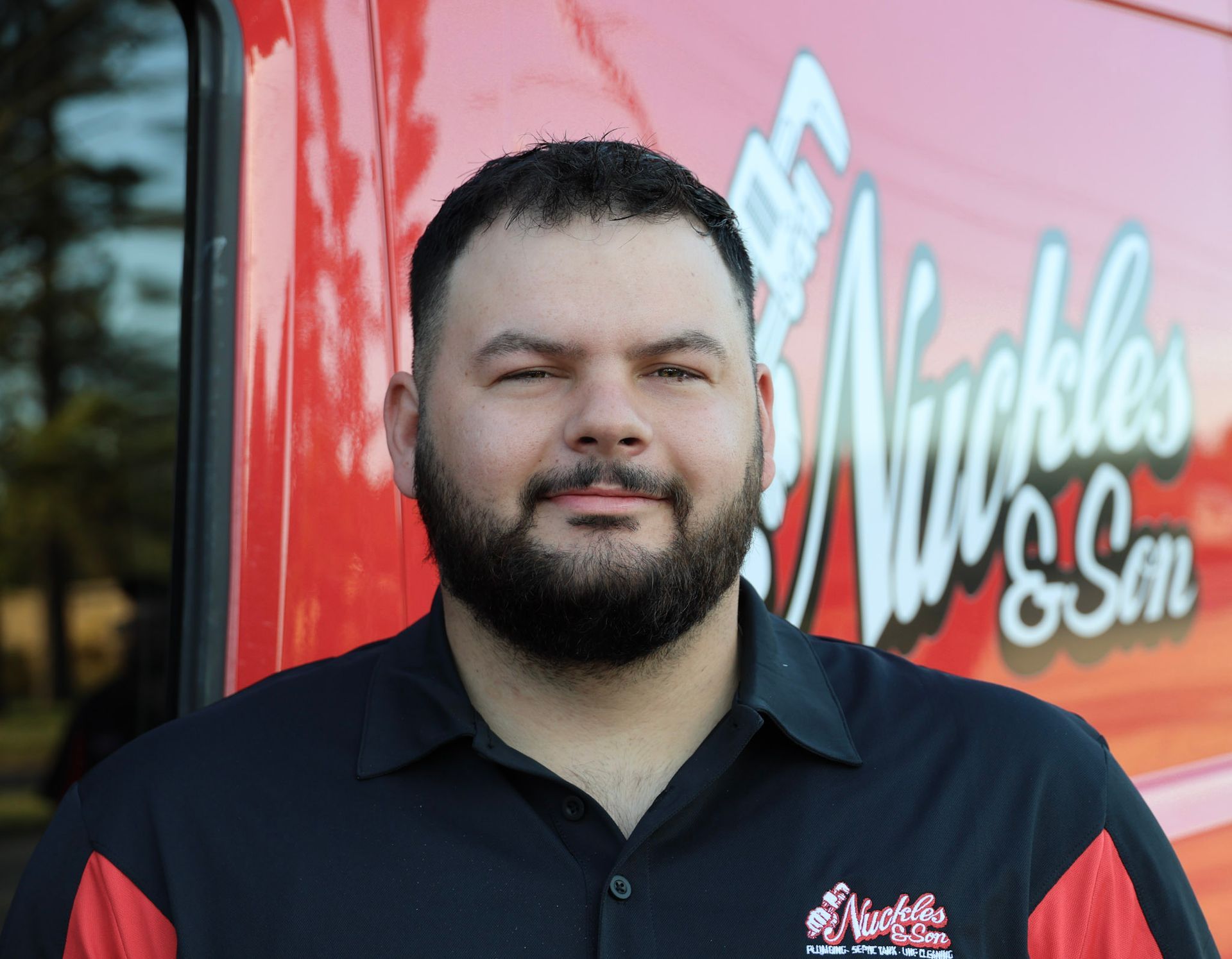 Portrait of a bearded man in a black and red polo standing before a red Muckles signage backdrop