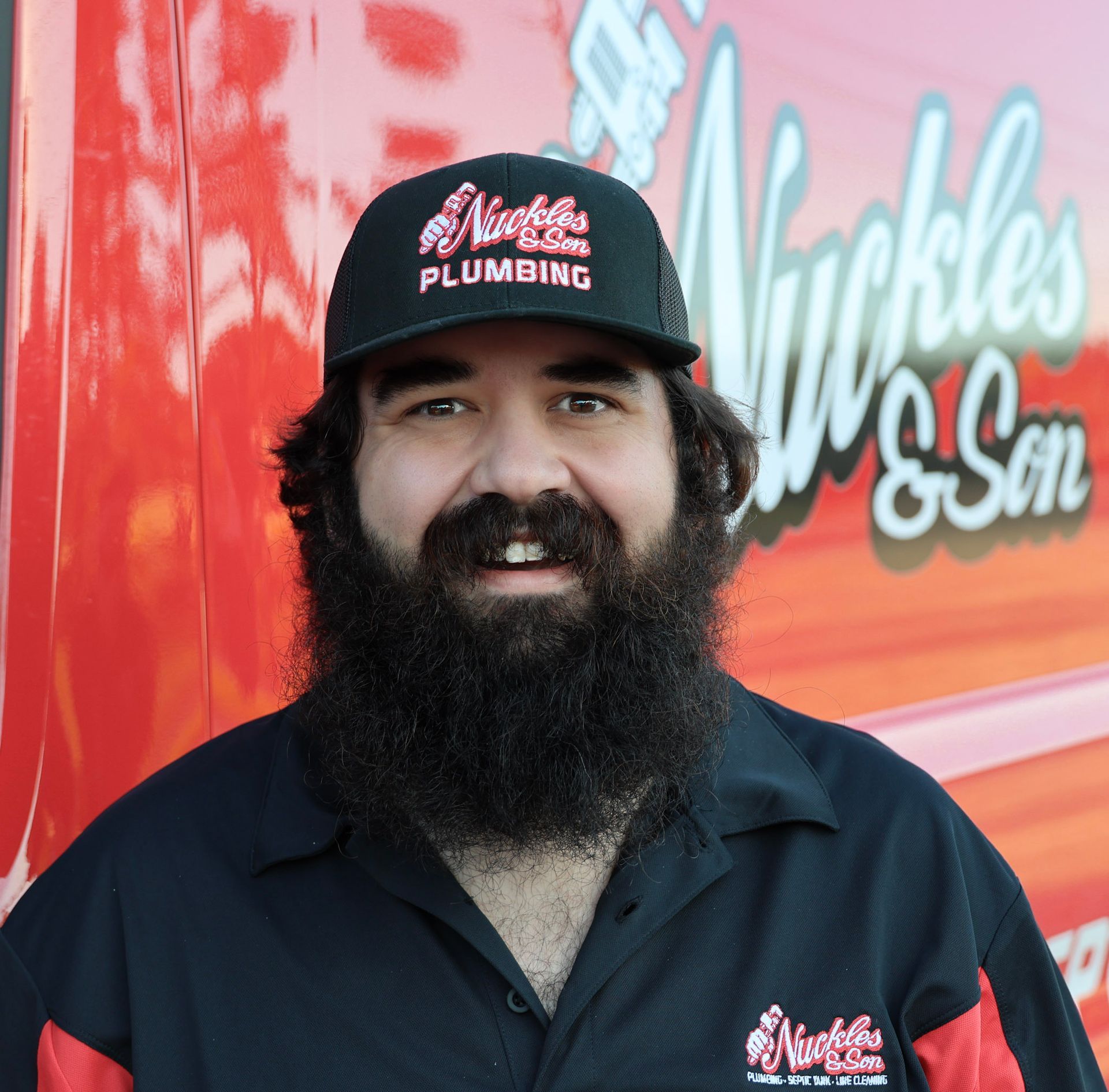 Man in black plumbing shirt and cap smiling in front of a red service truck