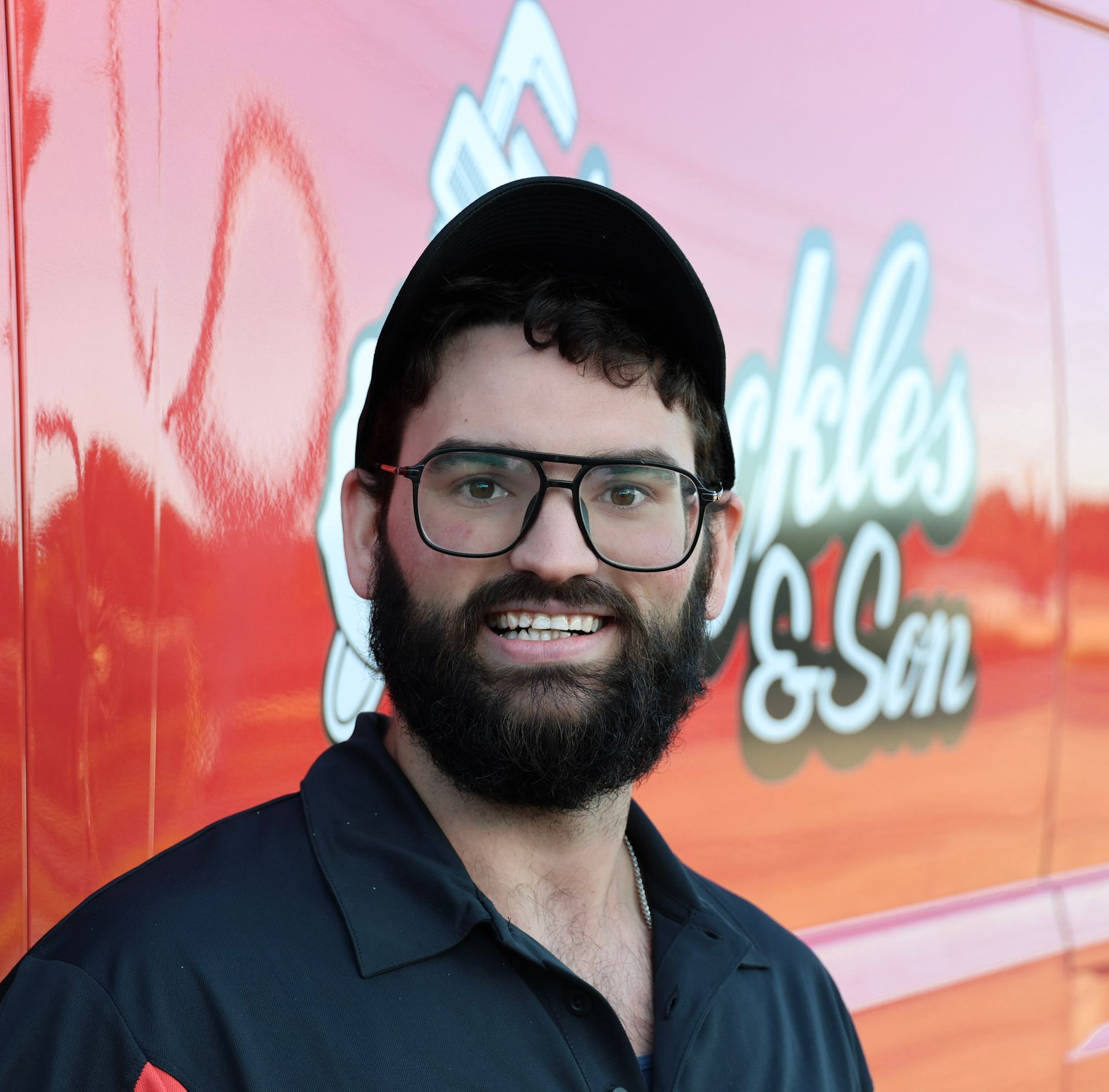 Smiling bearded man in glasses and a cap in front of a red-orange sign.