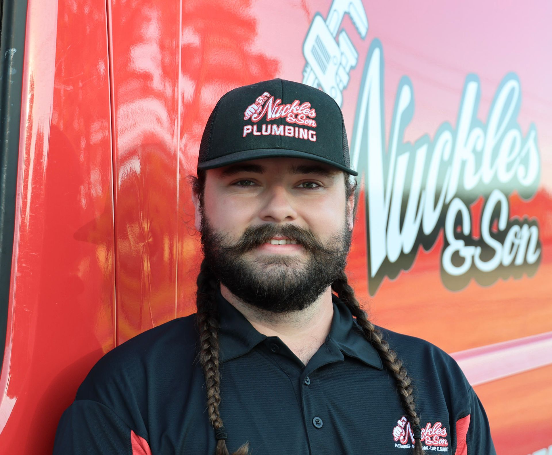 Bearded plumber in black shirt and cap standing by a red truck with “Nickles & Son” logo