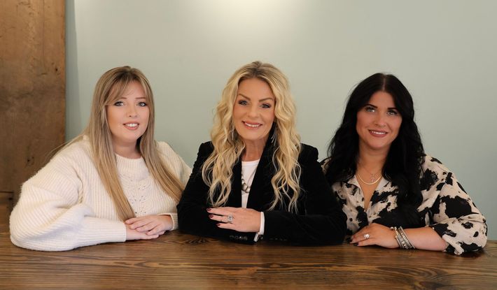 Three women seated behind a wooden table, posing indoors against a plain wall.