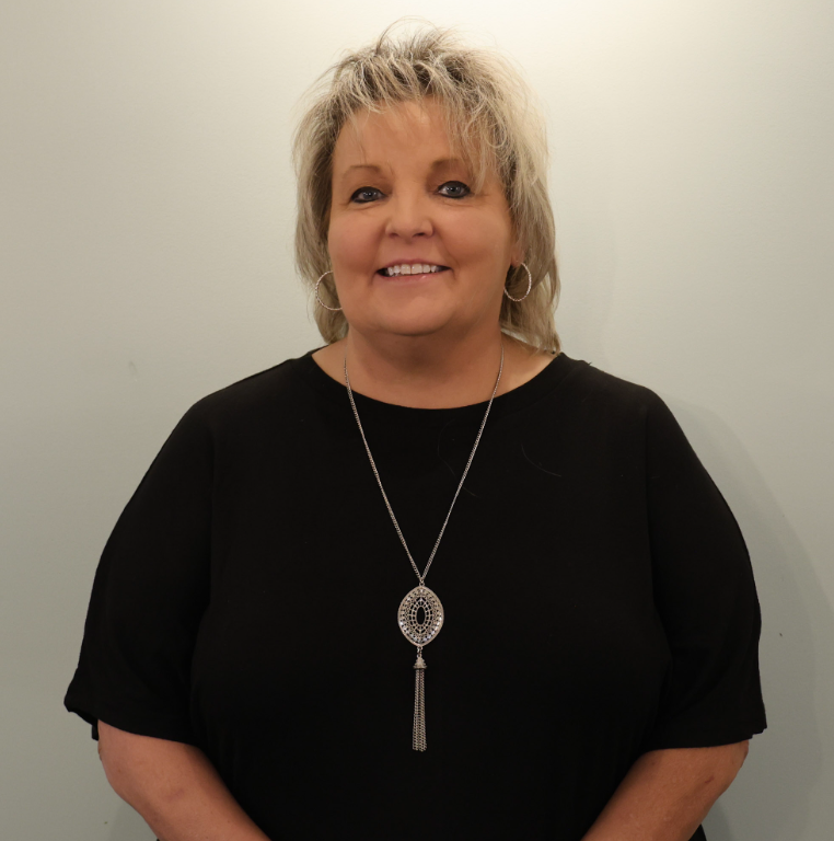 Woman smiling in a black top with a silver pendant necklace against a plain light background