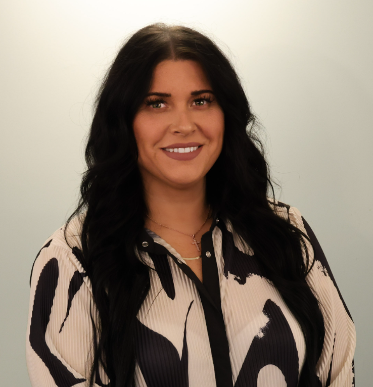 Smiling woman with long dark hair in a black-and-cream patterned blouse against a light background