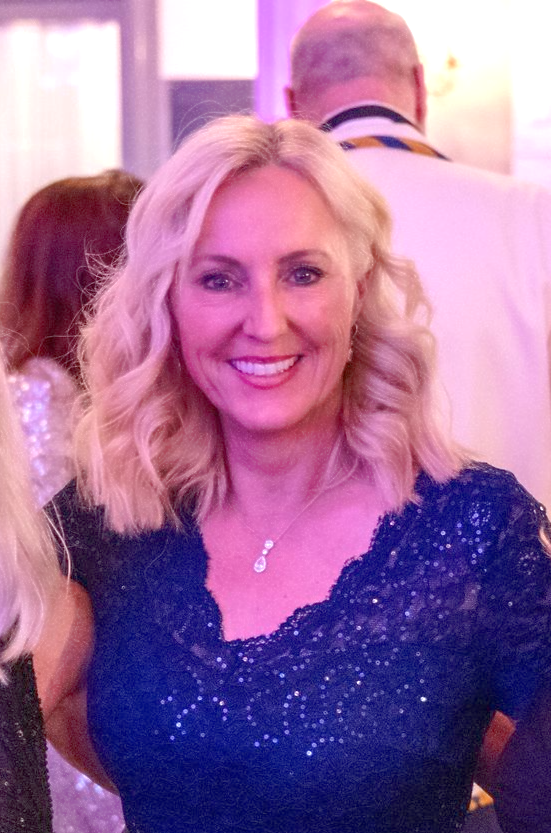 A woman with blonde hair is wearing a blue shirt and a necklace.