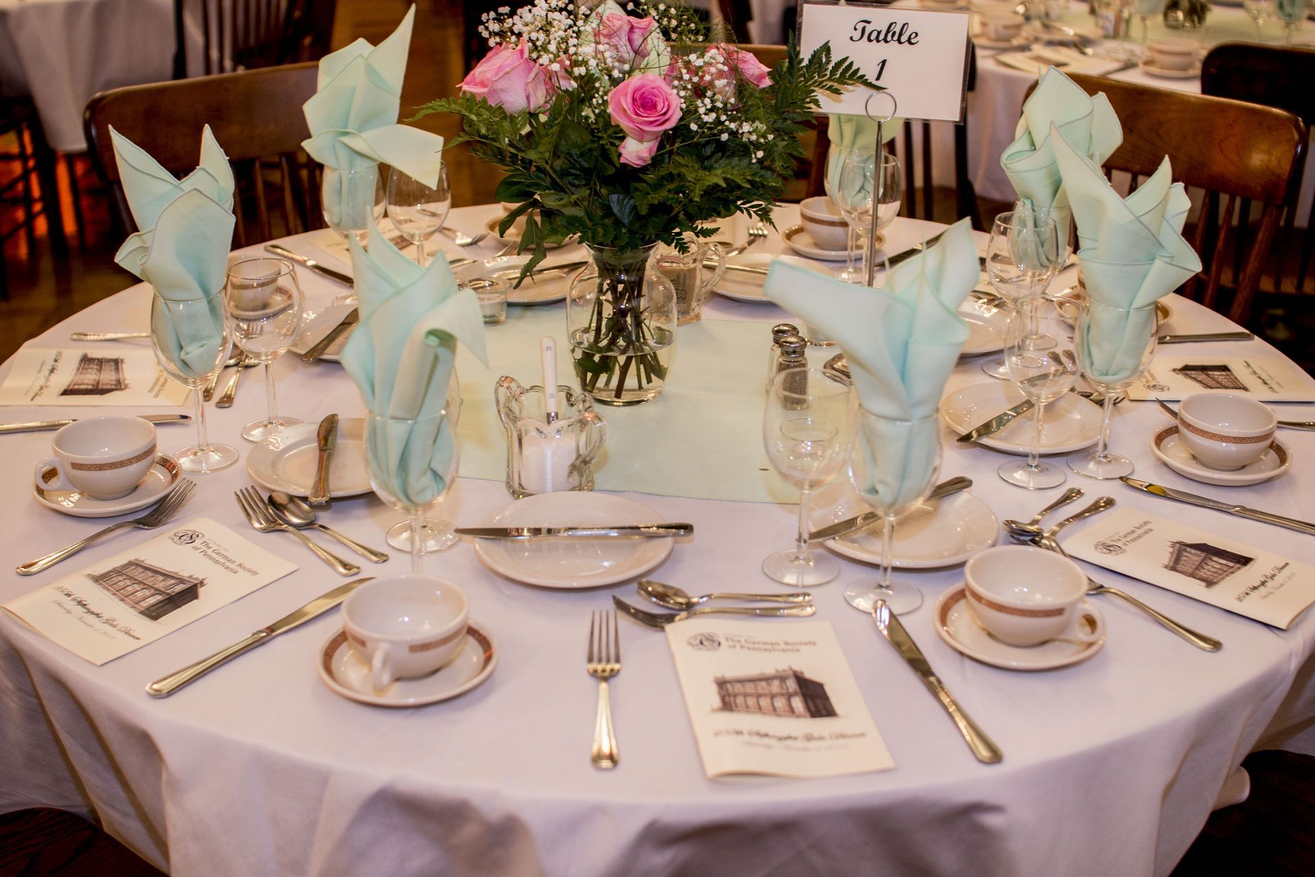 Elegantly set round banquet table with white linens, pale blue napkins, tea cups, and a pink floral centerpiece