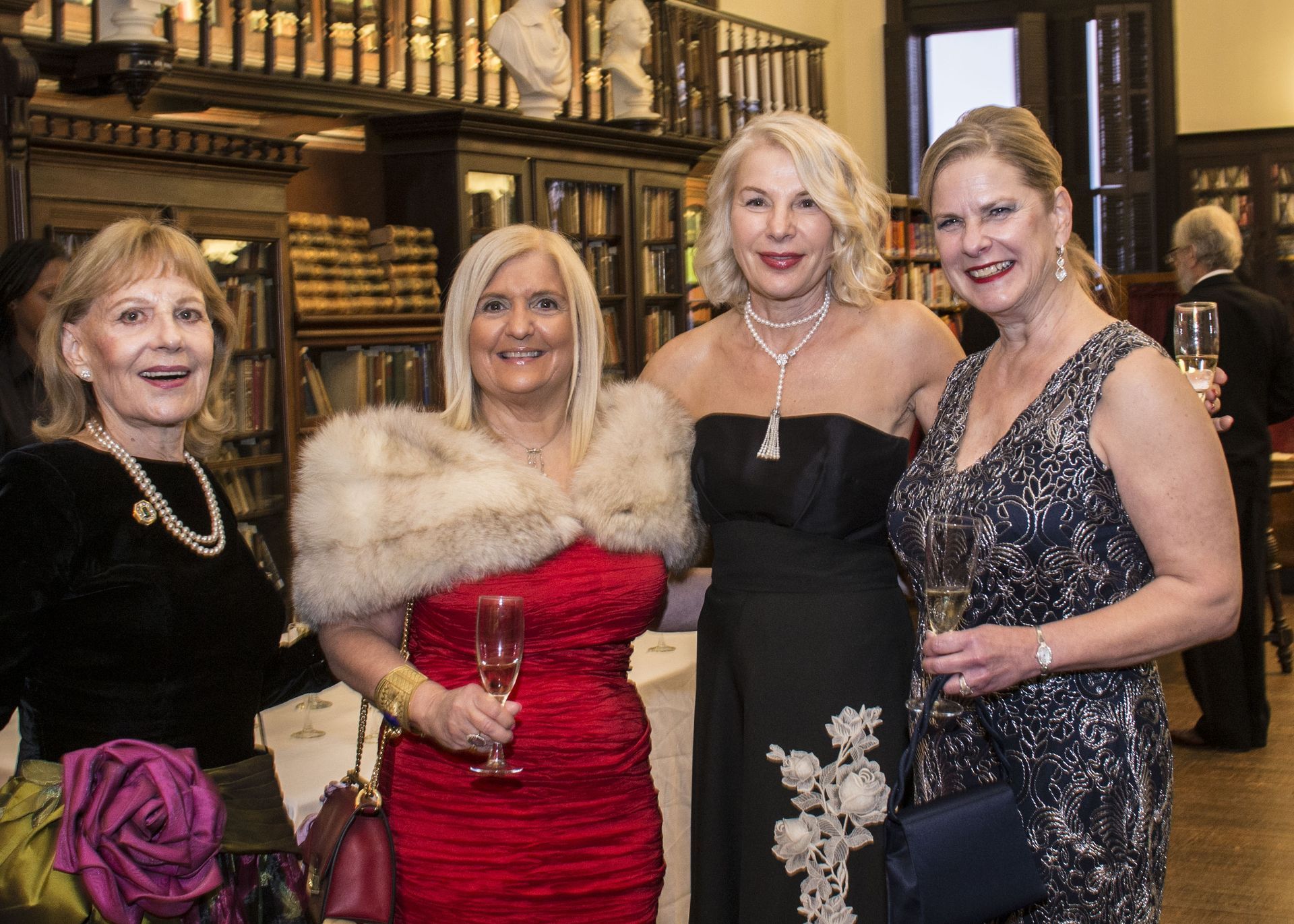 Four women in formal dresses posing together at an indoor event, one in a red gown with a fur wrap.