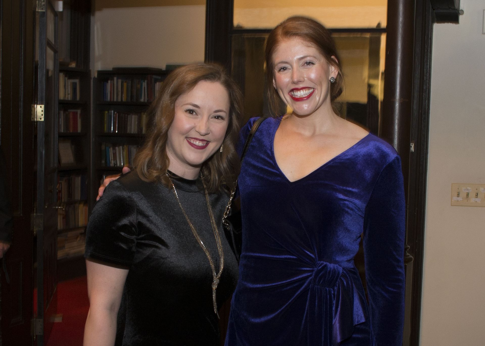 Two people smiling indoors, one in a black dress and one in a blue velvet dress.