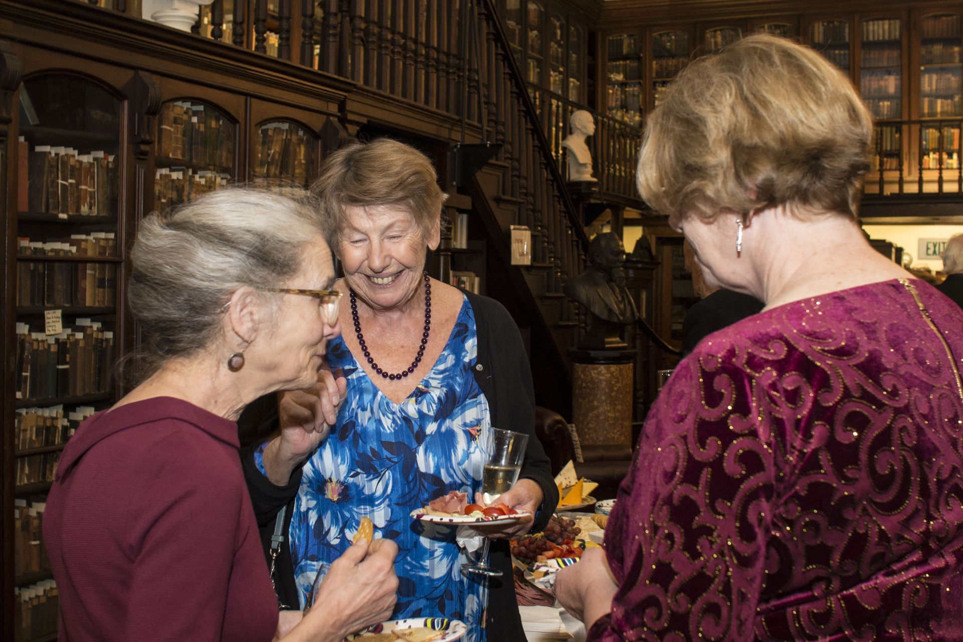 Three women chatting in a warmly lit library, one holding a plate of dessert