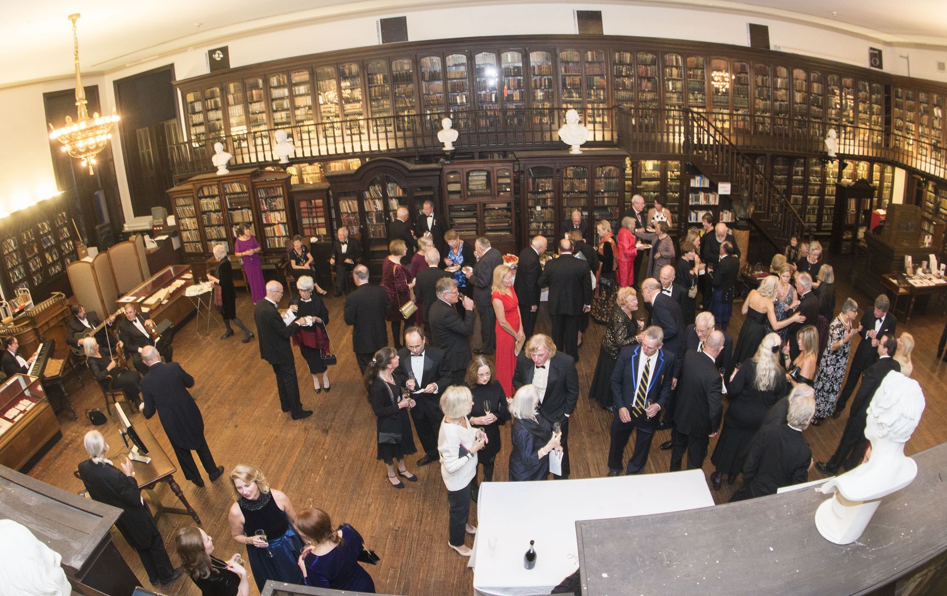 Crowded indoor event in a grand hall, with people mingling beneath ornate wall shelving and a white bust in front.