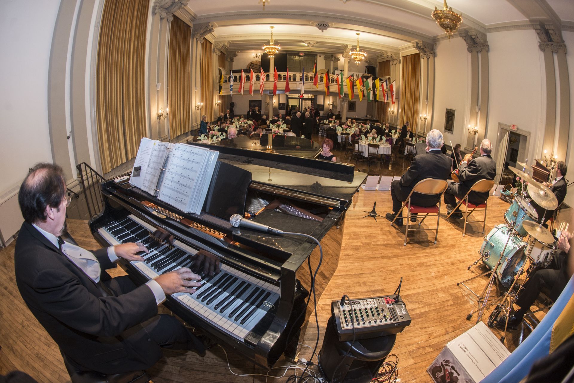 Musicians performing on stage in a formal concert hall, with keyboard and drum set in the foreground.