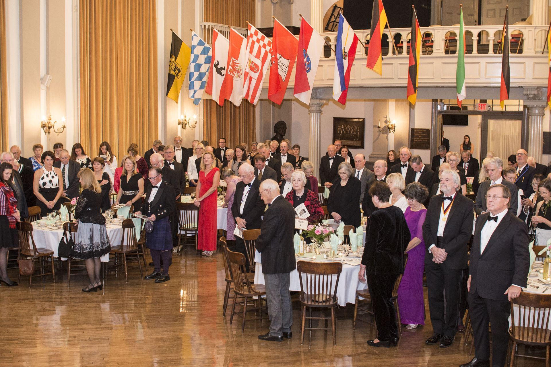 Formal indoor reception with guests in suits and dresses under hanging flags, applauding and chatting at round tables