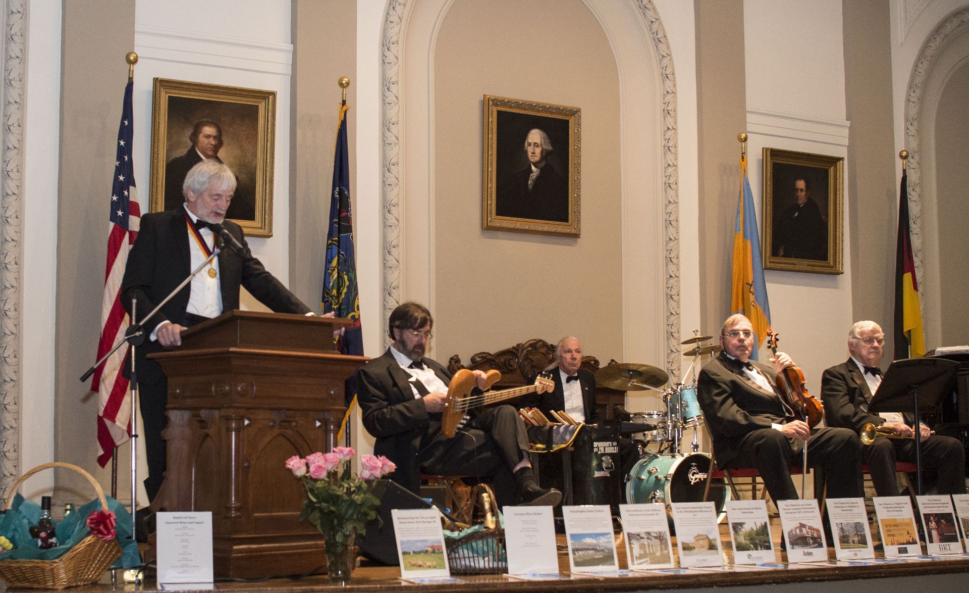 Speaker at podium in a formal hall with seated panelists and portraits on the wall