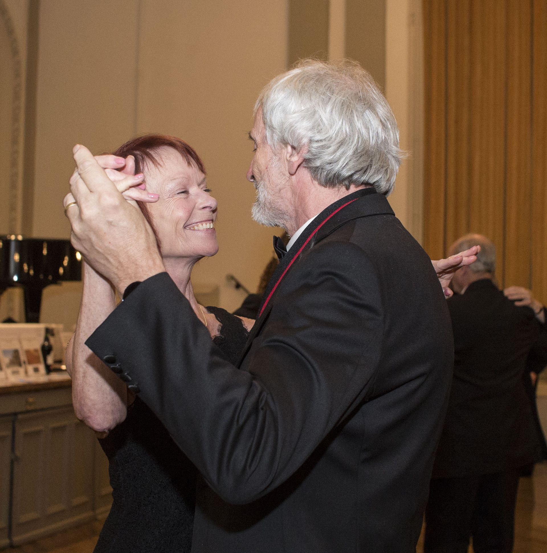 Two people dance closely at a formal indoor event, with one smiling and the other in a dark suit.