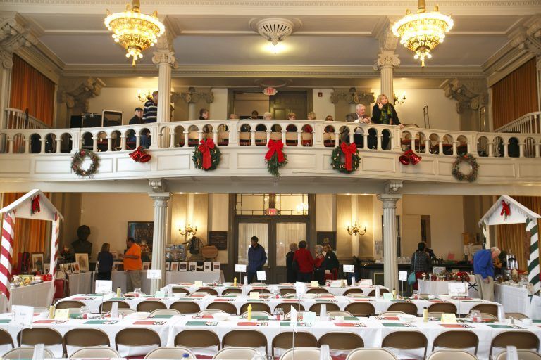 Decorated banquet hall with rows of white chairs, balcony guests, and chandeliers above the stage