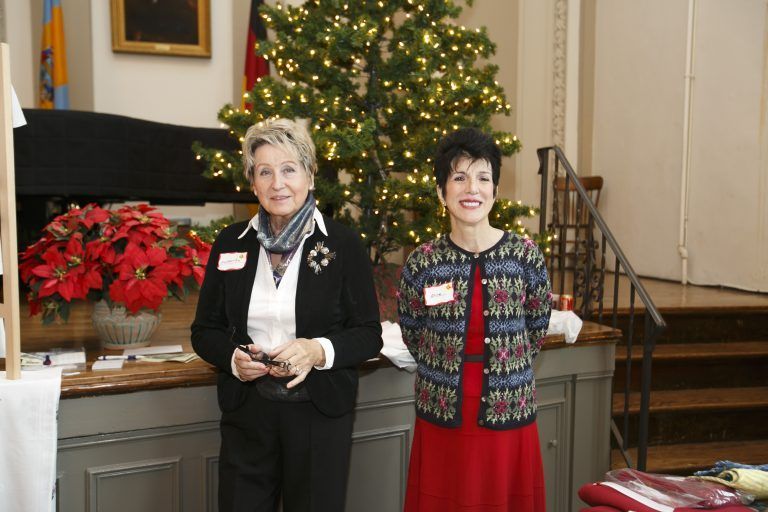 Two women standing indoors near a decorated Christmas tree and poinsettias, smiling at the camera