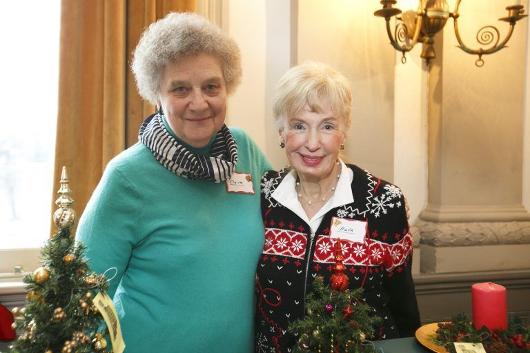 Two women pose indoors beside a decorated Christmas table and candles.