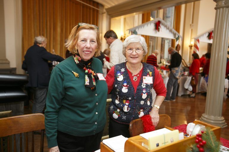 Two women smiling at a festive indoor gathering, with holiday decorations and tables behind them