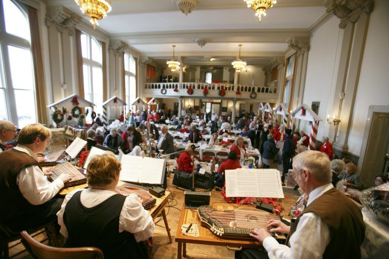 Orchestra performing in a grand concert hall with musicians seated under tall windows and ornate chandeliers