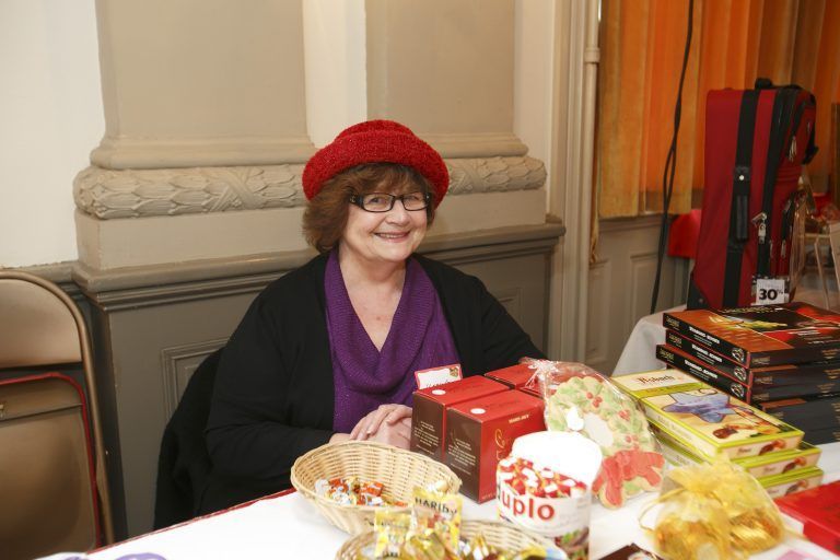 Woman in a red hat seated at a table with gift baskets and packaged goods at an indoor market.