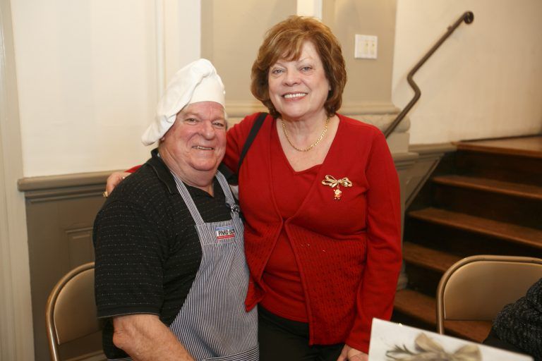 Two people smiling indoors near a staircase, one wearing a chef hat and apron, the other in a red sweater