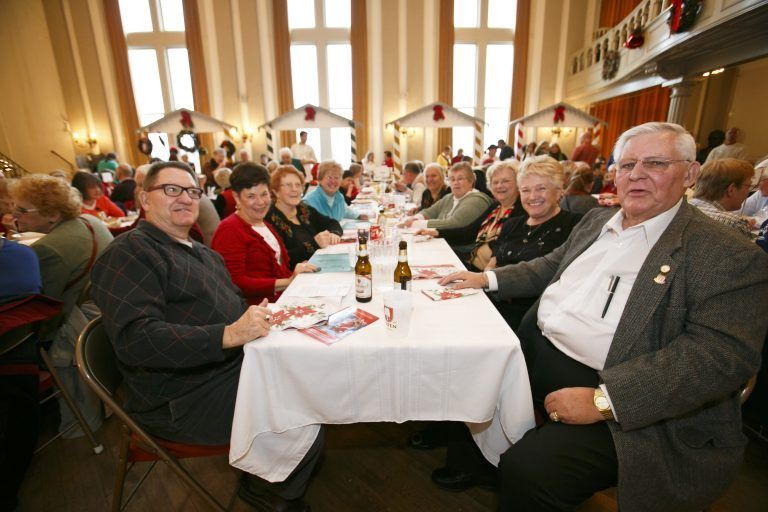 People seated at long banquet tables in a bright hall, talking and eating during a social gathering.
