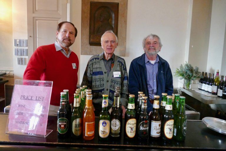 Three men stand behind a table of assorted beer bottles in a bar or restaurant.