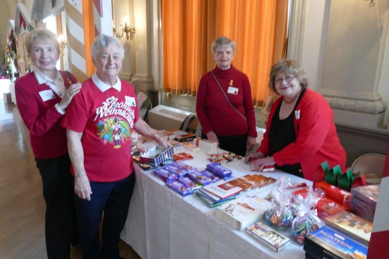 Four people stand behind a table of wrapped gifts and candy in a bright indoor room.
