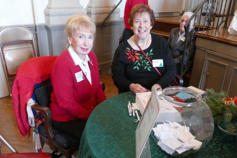 Two women seated at a table with a donation bowl and papers, smiling indoors at an event