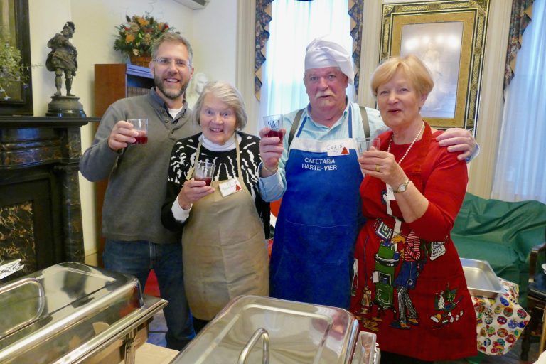 Four people in aprons and festive clothes raise glasses in a kitchen, smiling at the camera.