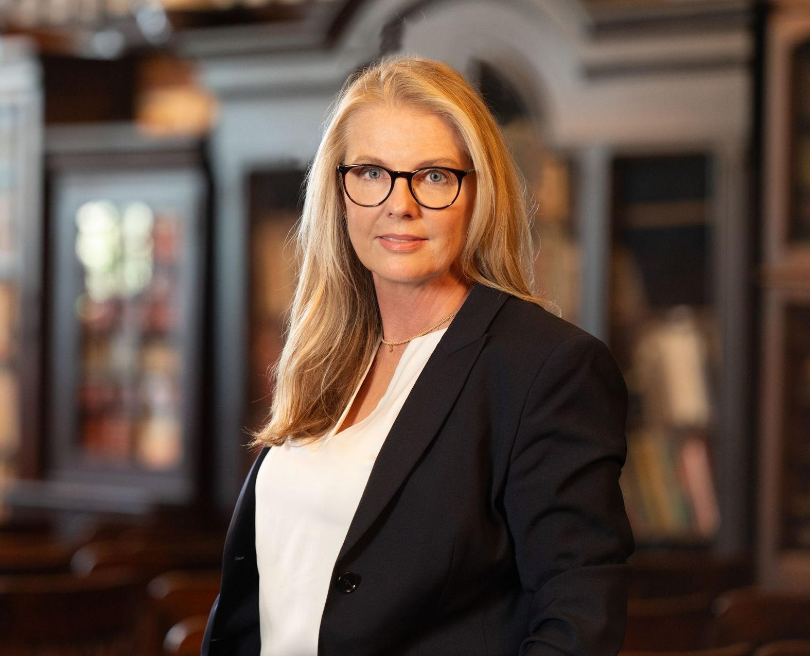 A woman in a suit and glasses is standing in a library.