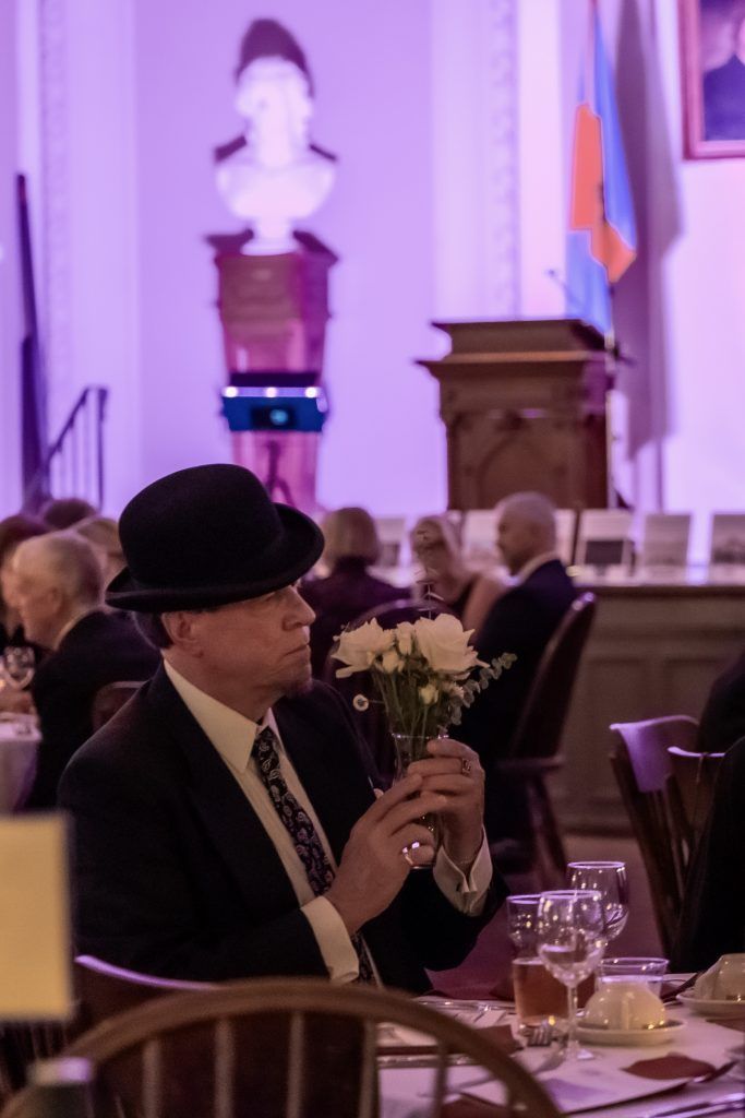 Man in a black hat holding flowers at a formal banquet table with a stage and podium in the background