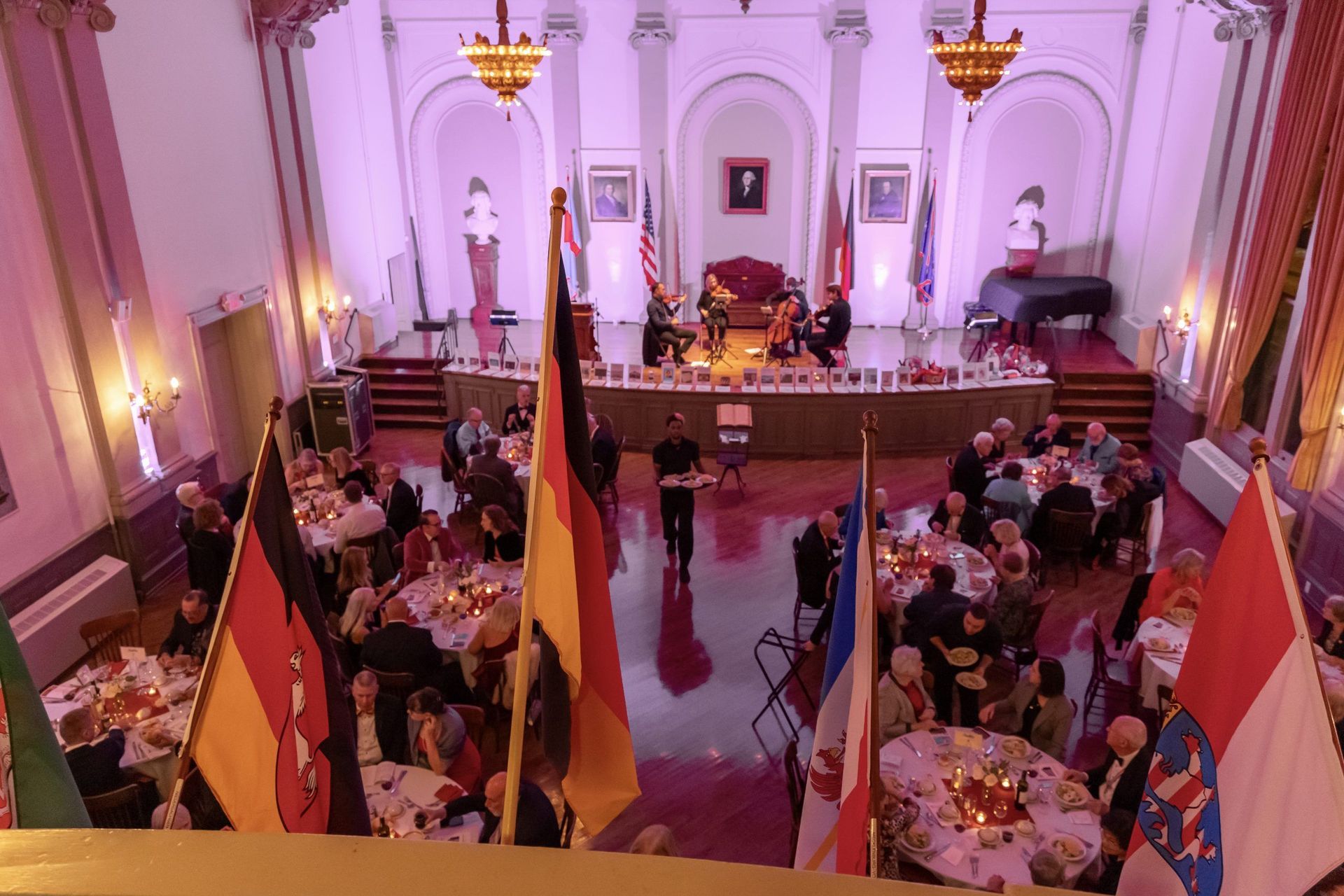 Formal banquet in a grand hall with round tables, flags, and guests seated at a stage-front event