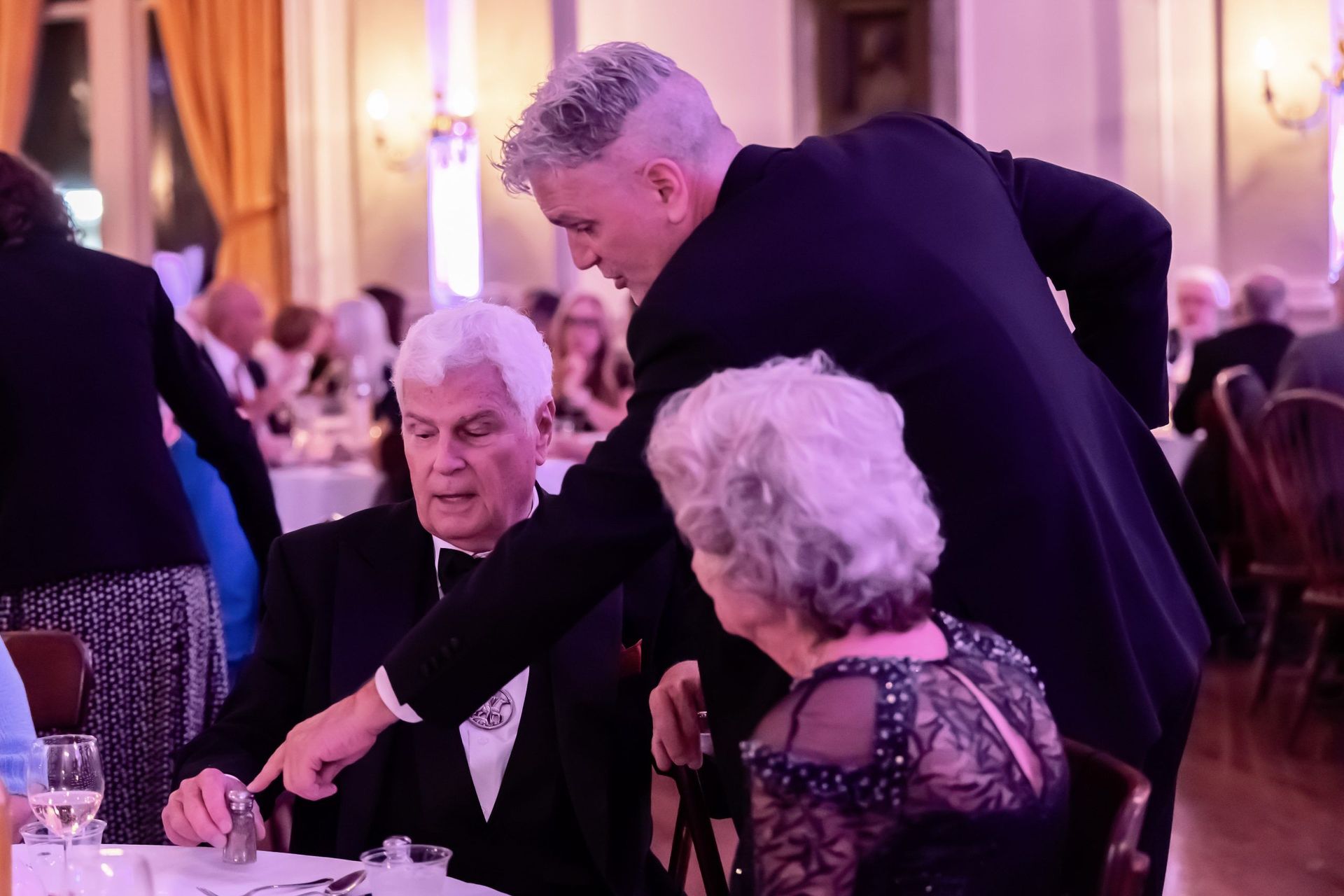 People seated at a formal event, with a man leaning over to speak to an elderly couple at a banquet table.