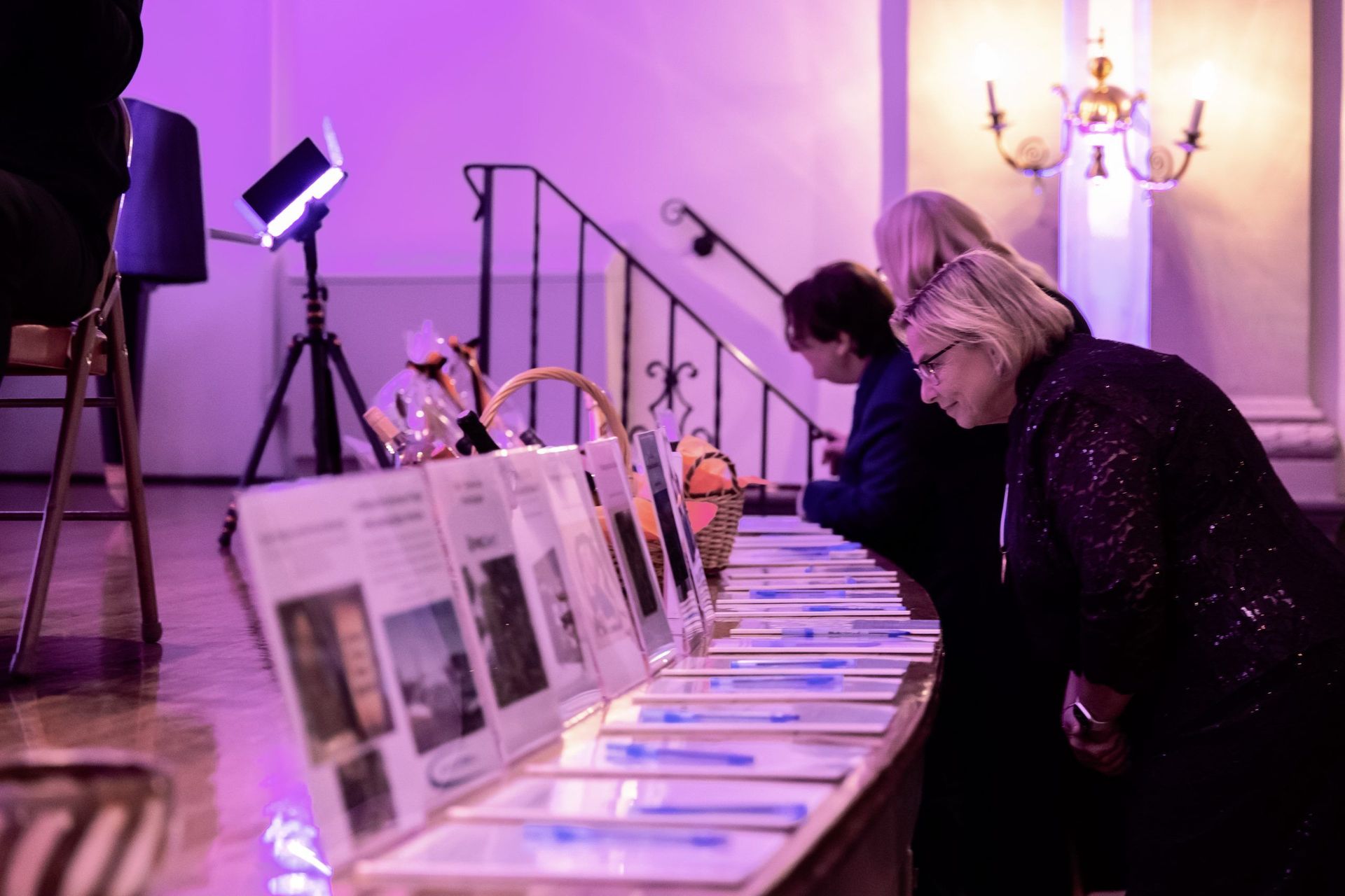People viewing brochures at a purple-lit exhibition table with framed displays and a tripod lamp