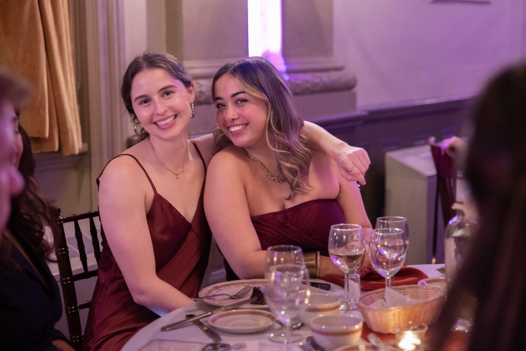 Two women in burgundy dresses smiling at a candlelit dinner table at a formal event