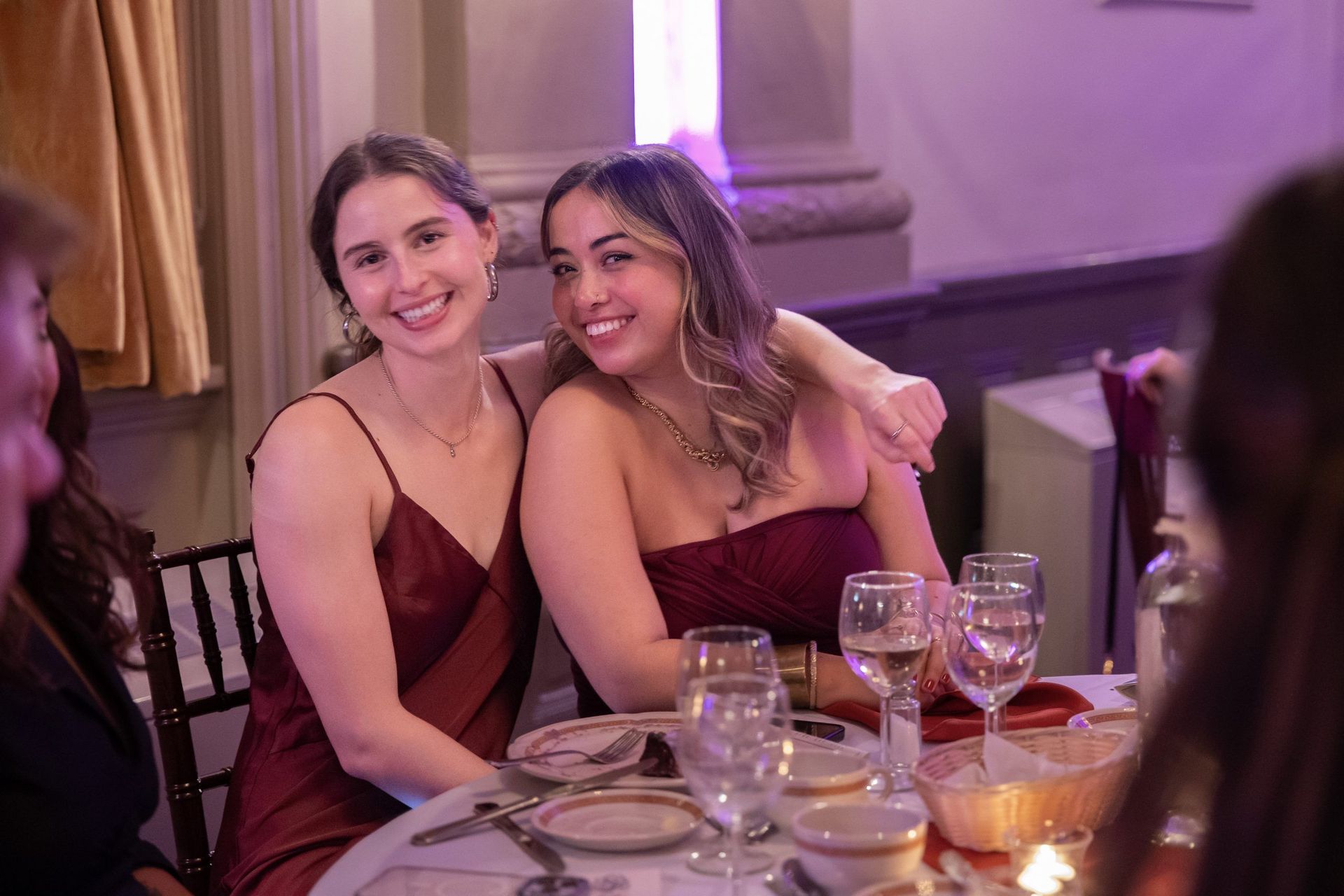 Two women in burgundy dresses smiling and hugging at a candlelit dinner table