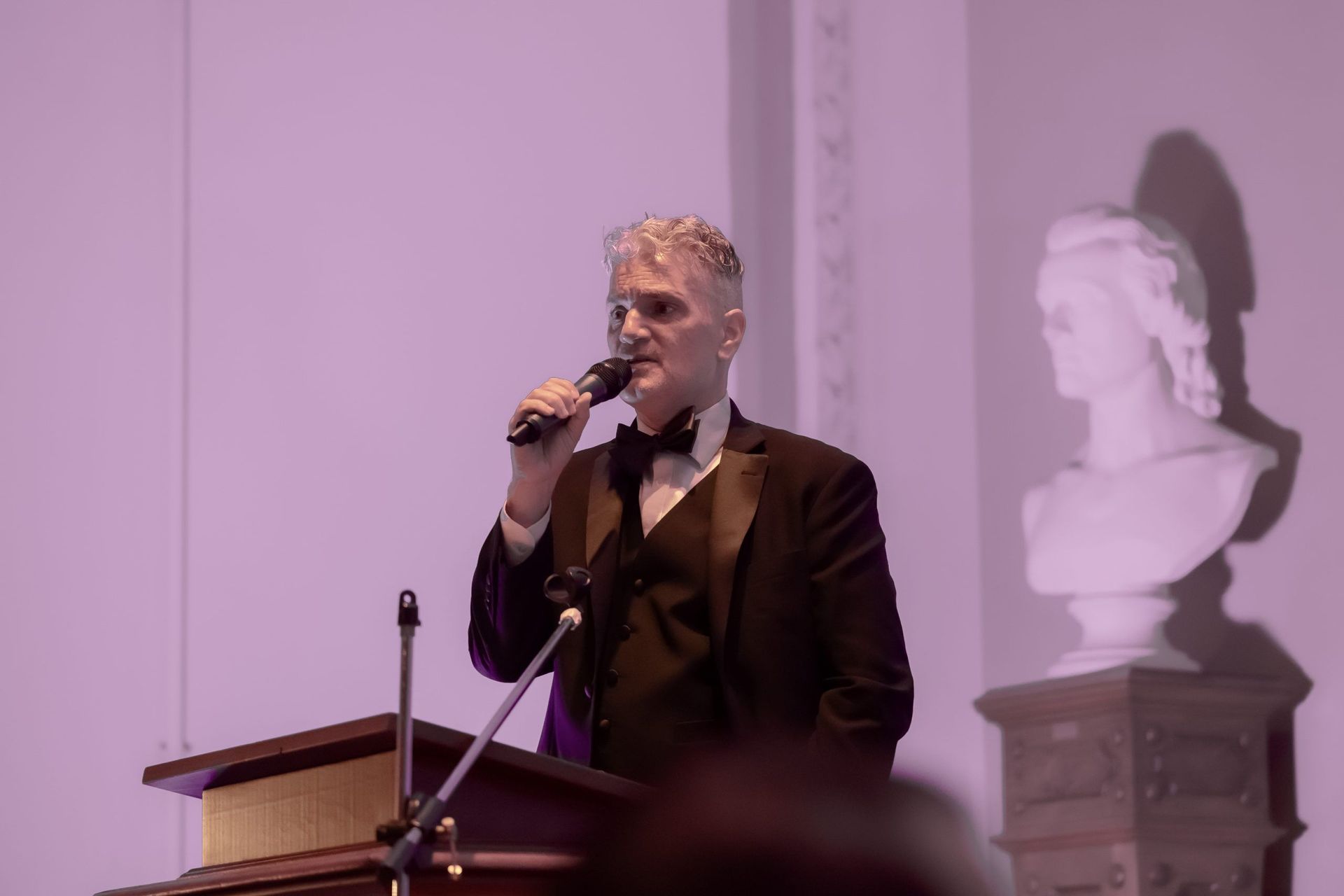 Man speaking into a microphone at a podium in a purple-lit room, with a bust statue in the background.