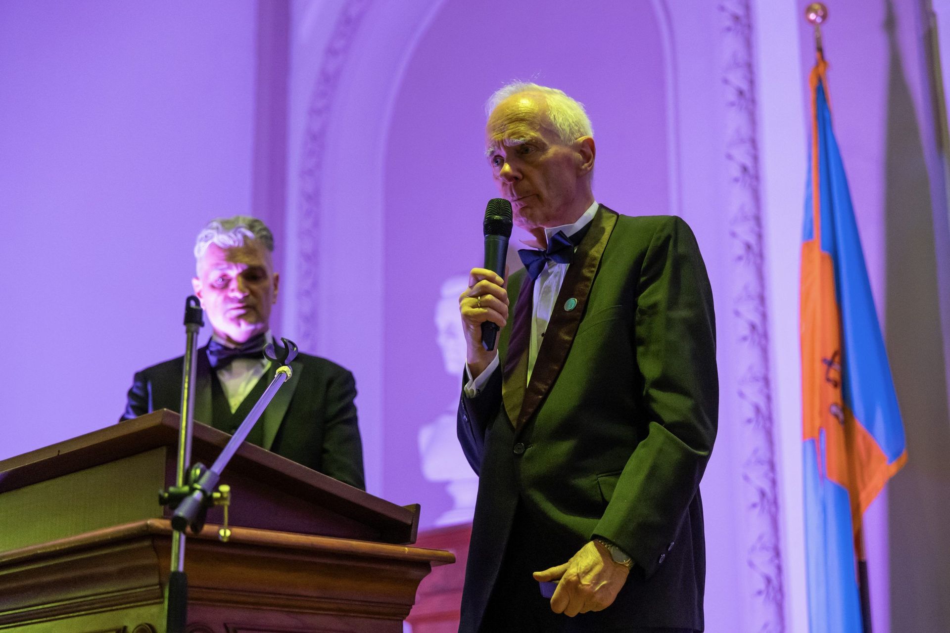 Two men at a podium in a purple-lit hall; one speaks into a microphone beside a flag.