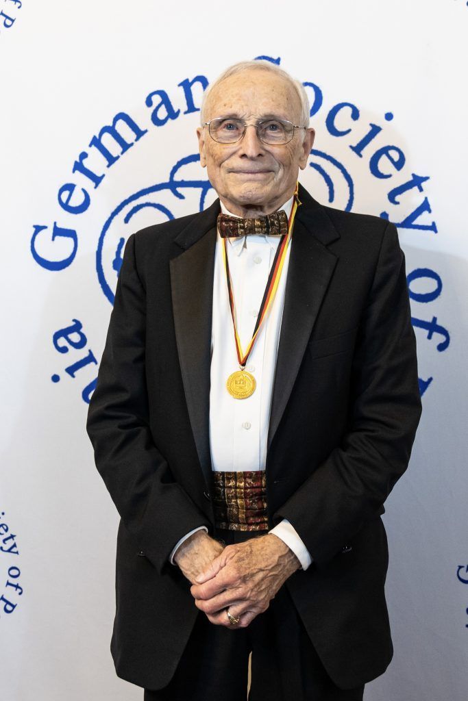 Man in black tuxedo with gold medal standing before German Society of title backdrop
