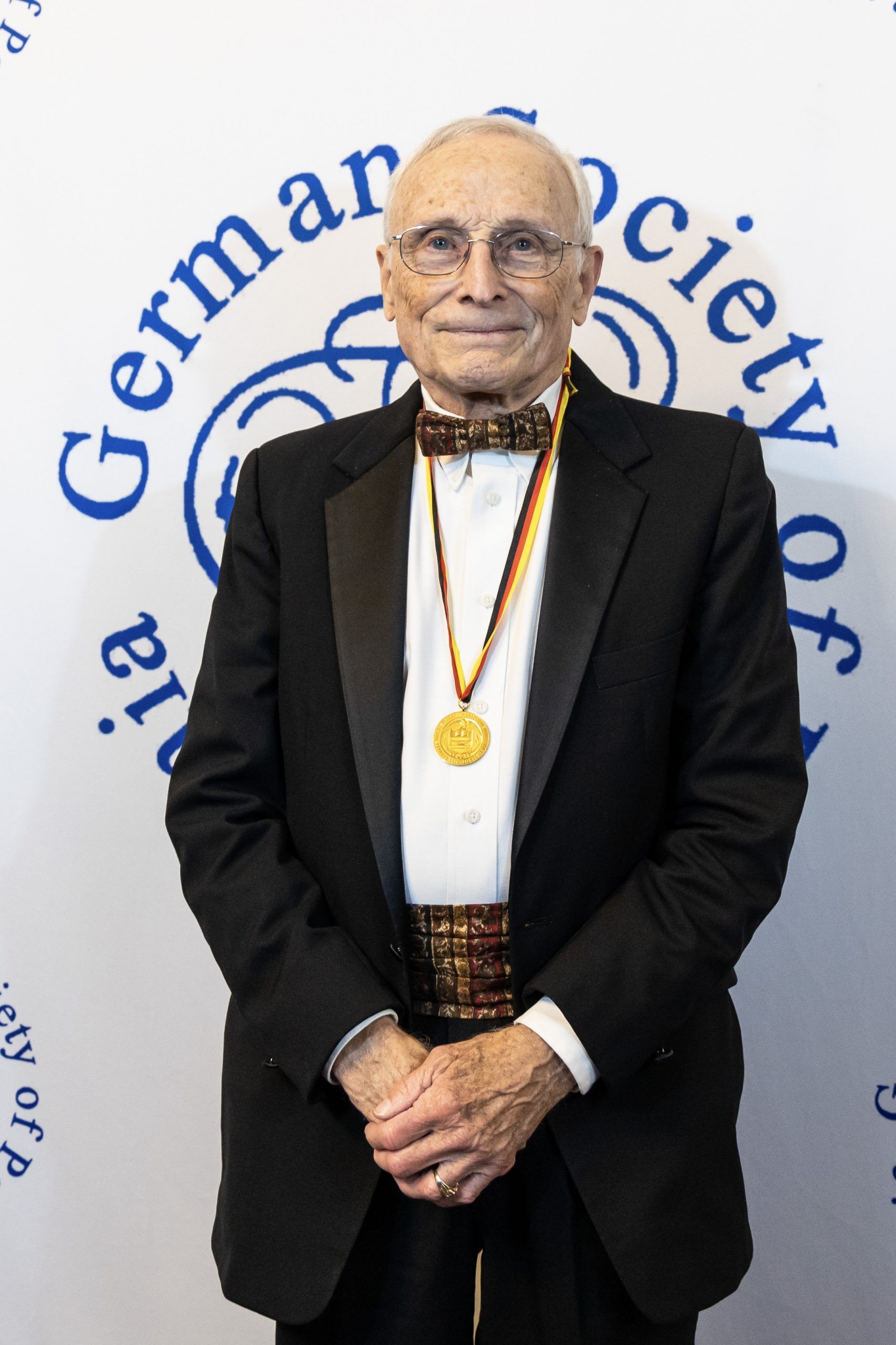 Elderly man in a black suit and gold medal standing before a German society backdrop.