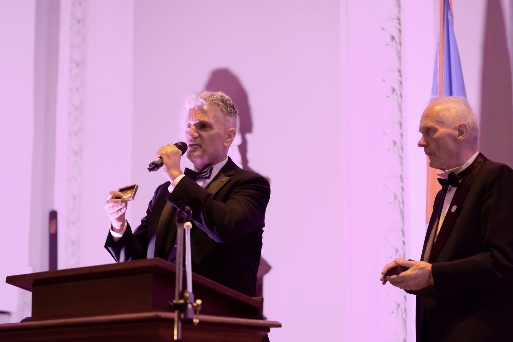 Two men at a podium speaking into microphones on a stage with purple lighting