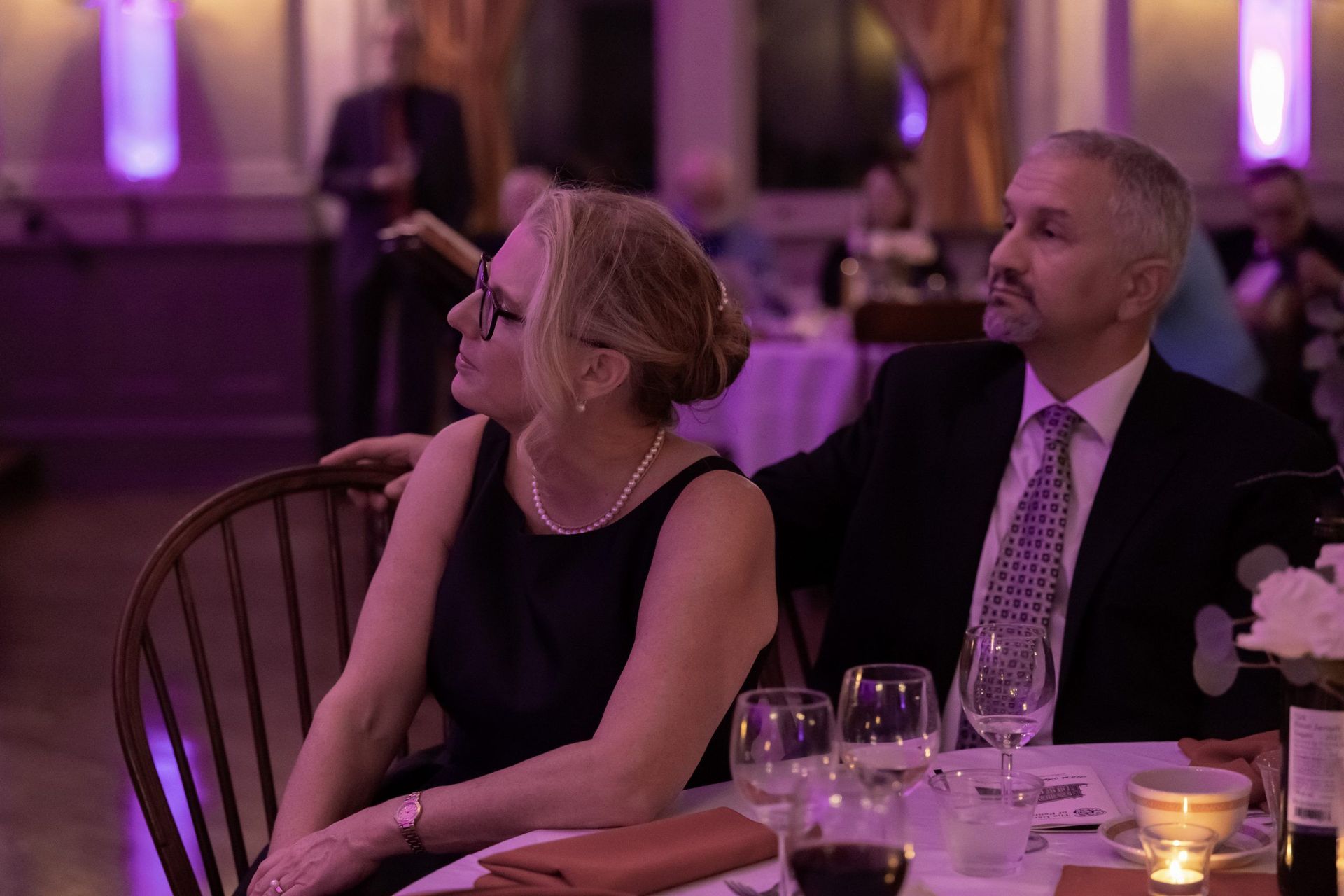 Two people seated at a candlelit banquet table, looking left in a purple-lit event hall.