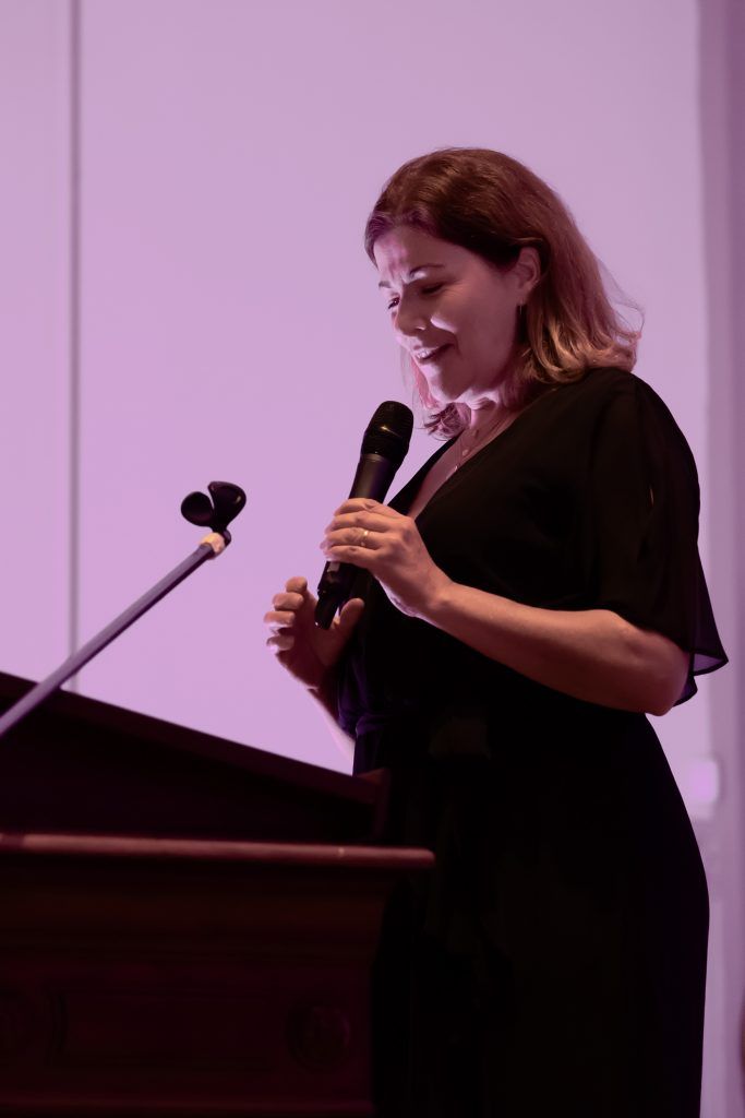 Woman speaking into a microphone at a podium in a dimly lit room with purple lighting