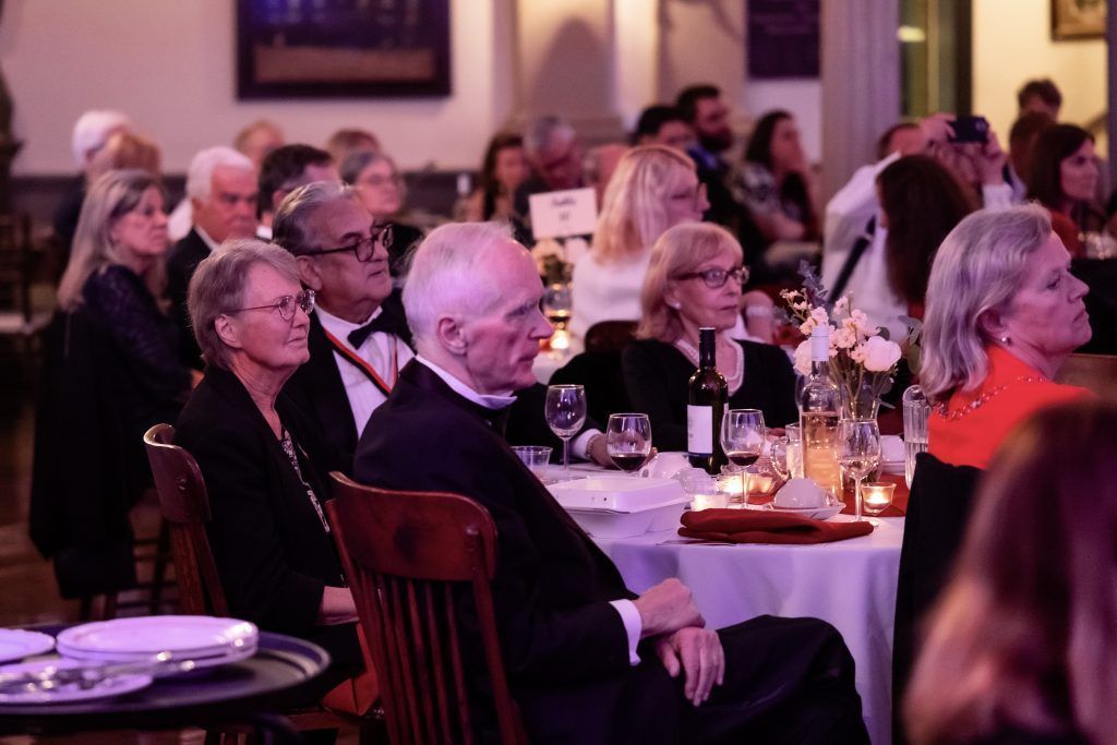 Audience seated at round tables in a warmly lit event hall, watching a presentation