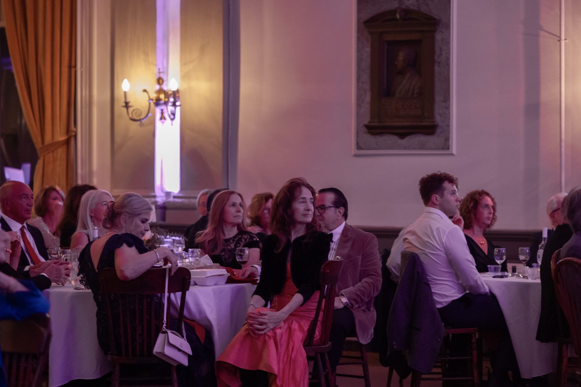 People seated at tables watching a formal event in a warmly lit hall