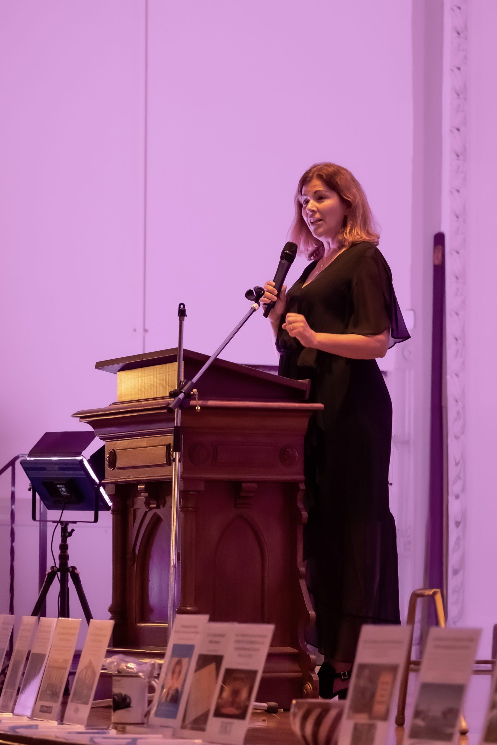 Woman speaking at a podium on a stage with purple lighting, in front of display tables