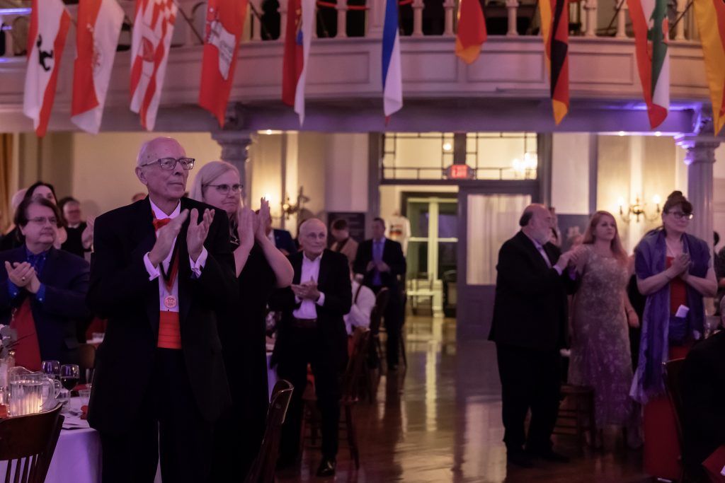 Guests applaud and dance at a formal indoor reception under hanging national flags.