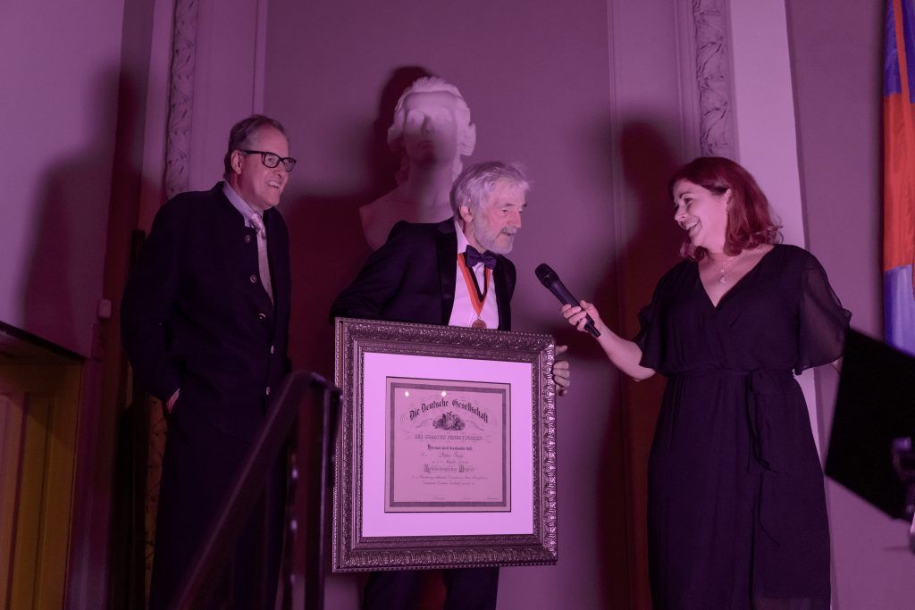 Three people stand onstage at a podium during an award presentation, with a microphone and framed plaque.
