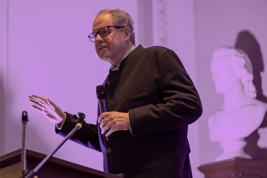 Speaker at a podium gesturing during a presentation, lit by purple stage lighting.